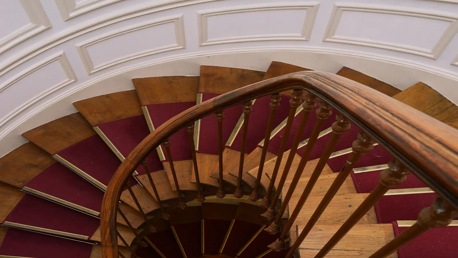 Escalier en colimaçon en bois avec rampe sculptée et marches recouvertes d’un tapis rouge.