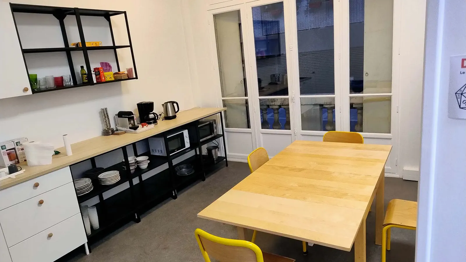 Espace cuisine et pause avec table en bois, chaises jaunes et équipements comme micro-ondes, cafetière et bouilloire.