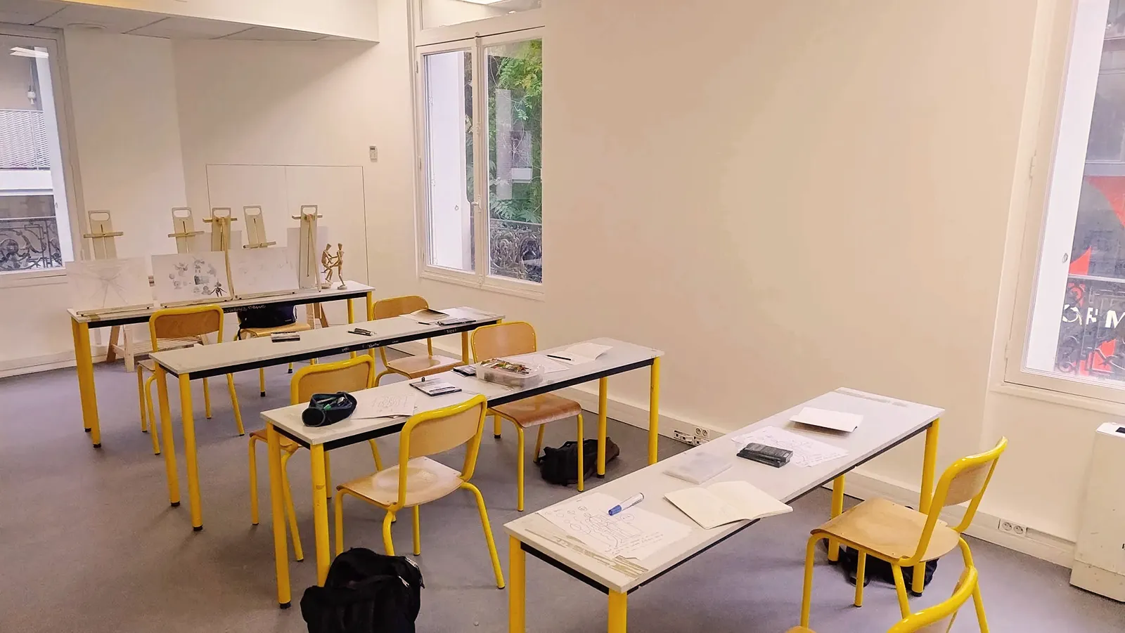 Salle de classe lumineuse avec tables et chaises jaunes, équipée pour un cours de dessin avec des chevalets et des croquis.