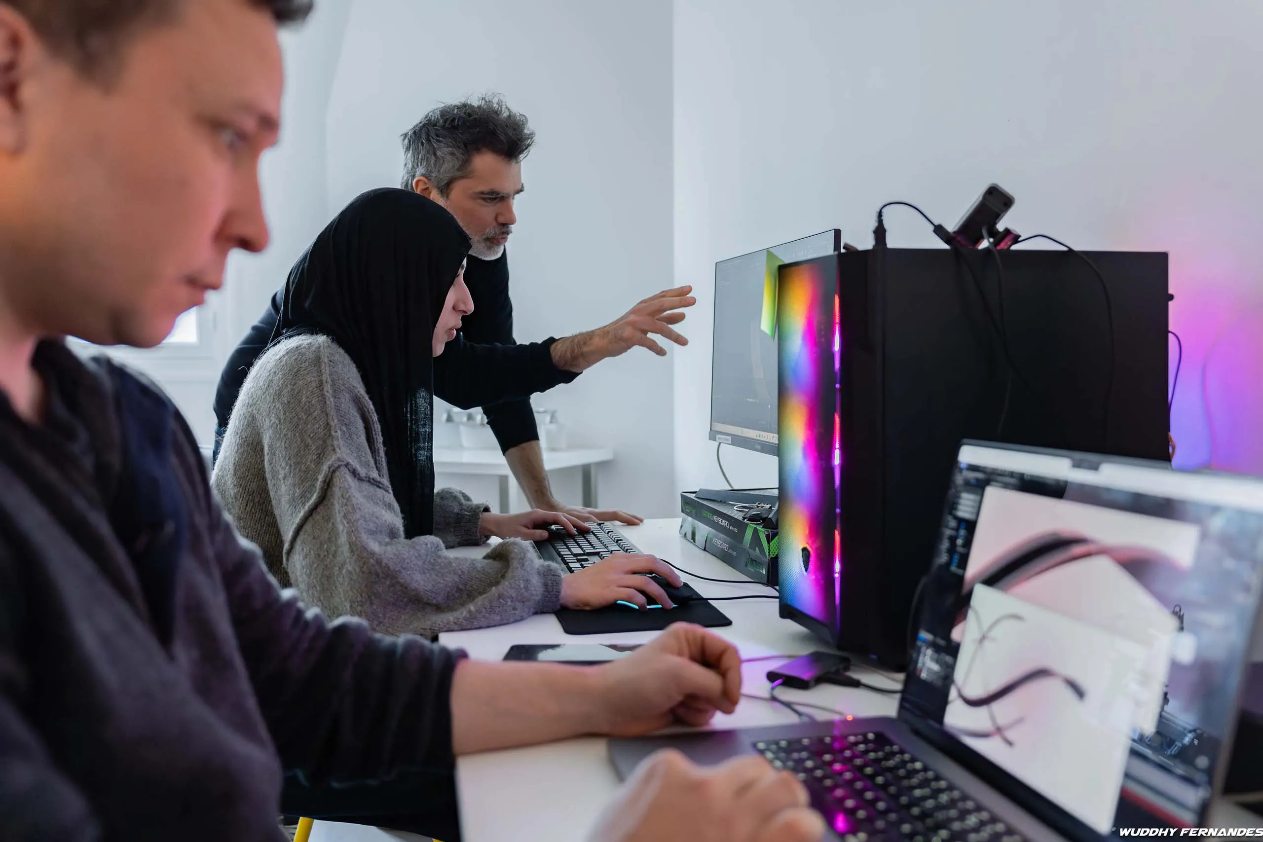Trois personnes travaillent sur des ordinateurs dans un espace de travail collaboratif, avec un formateur qui accompagne une participante devant un ordinateur.